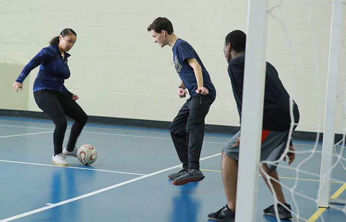 a woman kicking a soccer ball as two men play defense and goalie